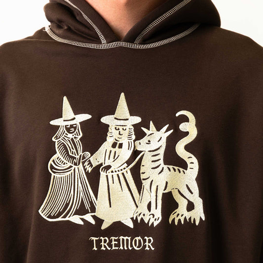 Brown hoodie with white graphic design of two figures and a creature, labeled 'TREMOR'.