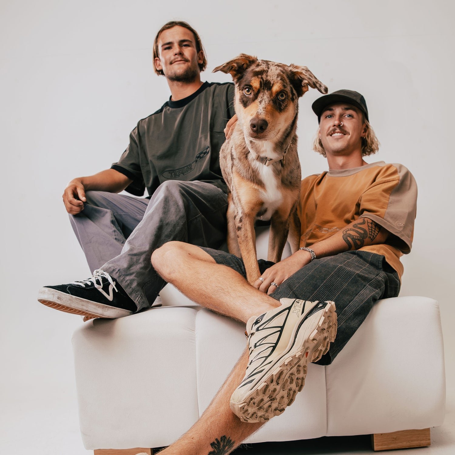 Two people sitting on a white couch with a dog between them, against a plain background.