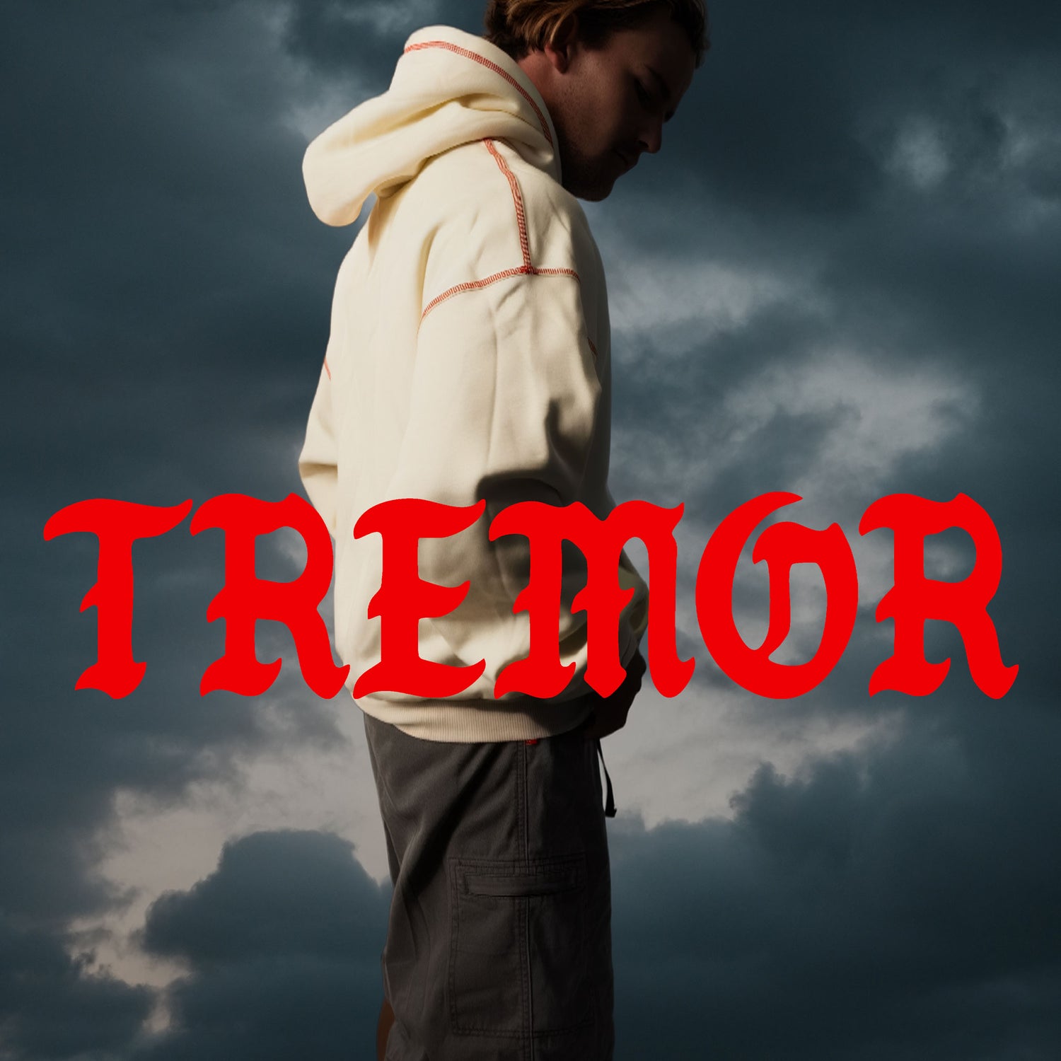 Person wearing a beige hoodie with red text 'TREMOR' against a dark cloudy sky