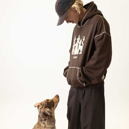 Person wearing a brown hoodie with a logo and text, standing next to a dog on a plain background